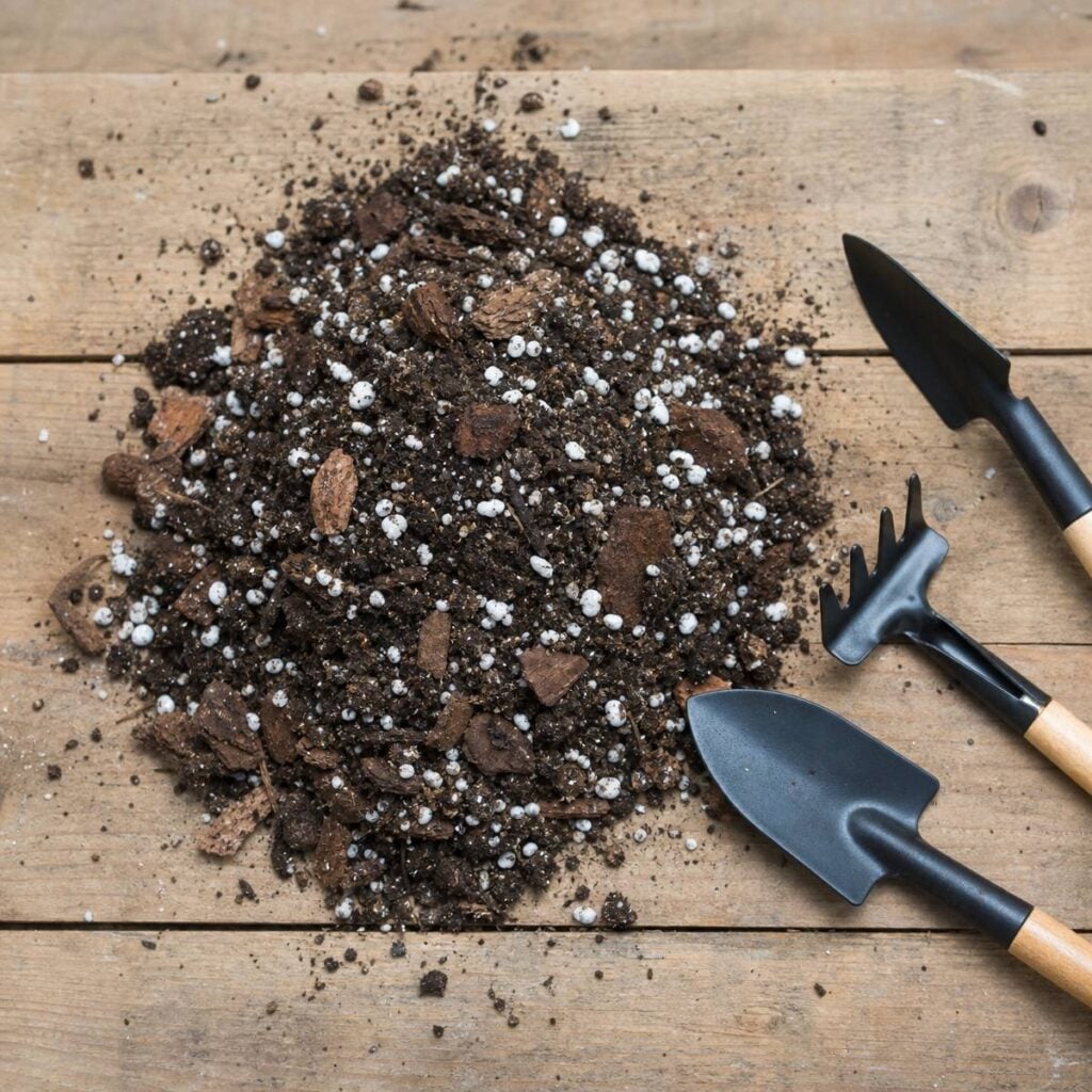 A mound of dark potting mix with white perlite granules and bark chunks sits on a wooden table beside two small trowels and a hand rake.