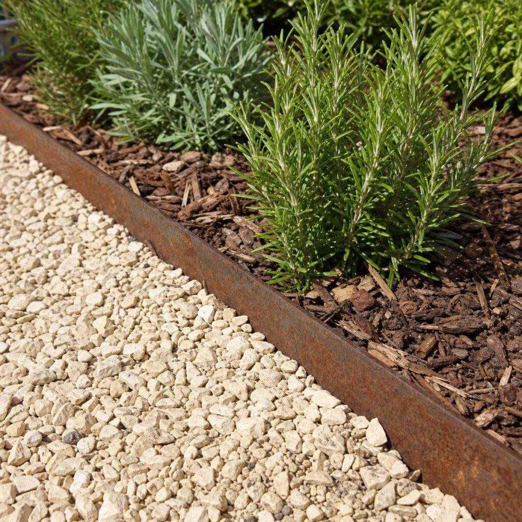 A rusted metal edging strip runs in a straight line separating pale gravel from a mulched bed with rosemary and lavender shrubs.