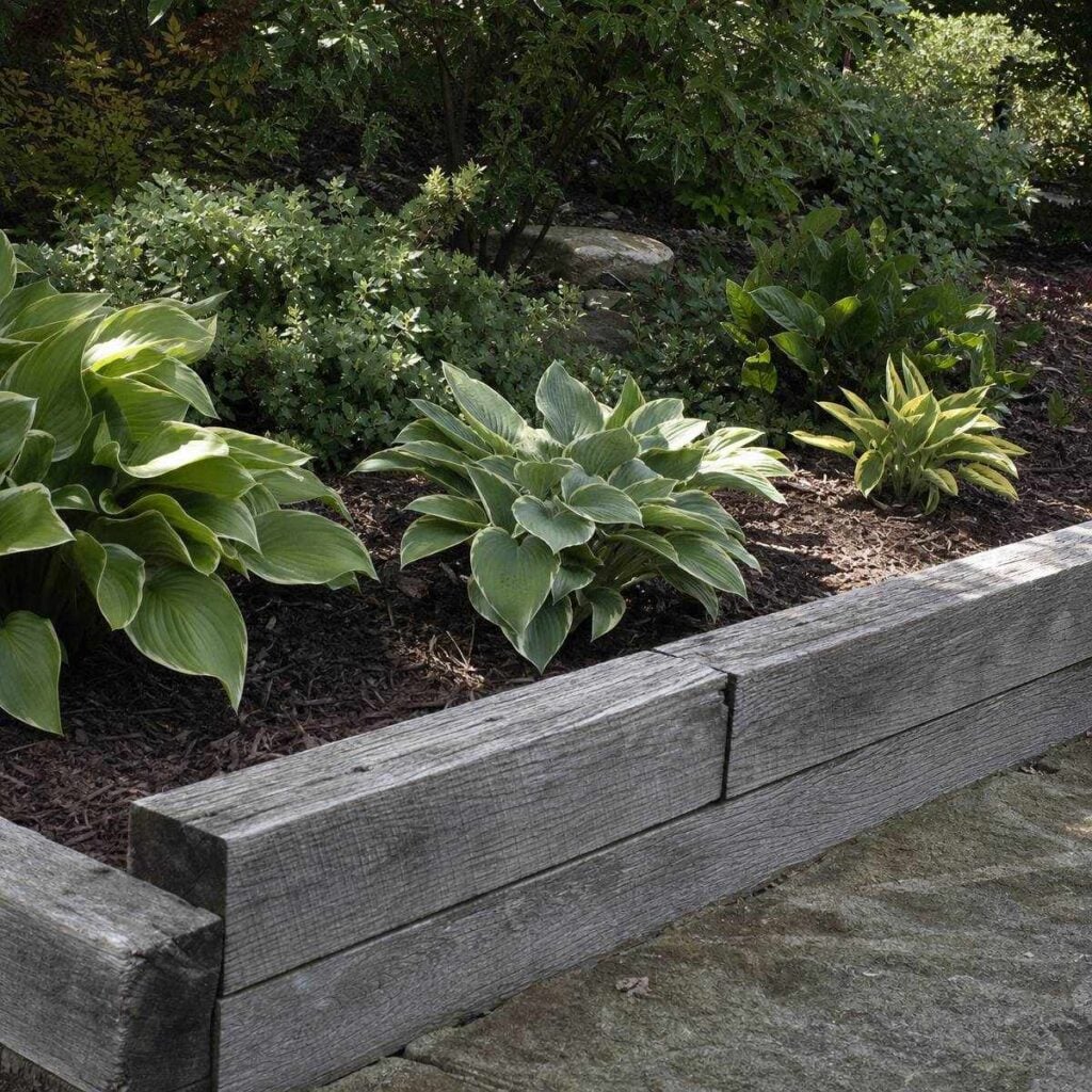 Weathered gray wooden timbers form a low border along a mulched bed of hostas and shrubs beside a stone path.
