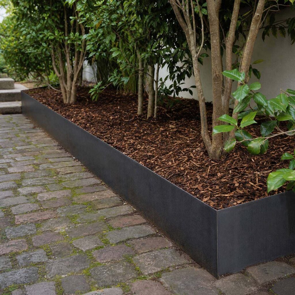 A dark metal-edged raised bed lines a mossy cobblestone path, filled with brown mulch and multi-stemmed shrubs against a white wall.