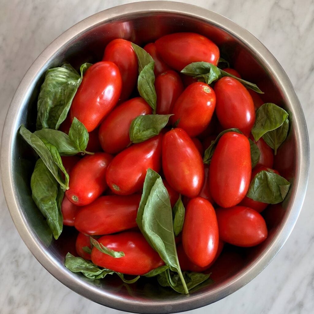 A stainless steel bowl viewed from above holds elongated red San Marzano tomatoes mixed with fresh green basil leaves on a marble countertop.